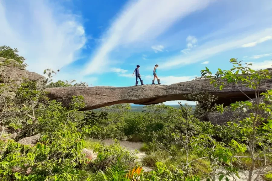 Two travellers cross a natural rock bridge in Chapada dos Guimarães, surrounded by native vegetation and a blue sky with clouds, in the heart of Mato Grosso, Brazil.