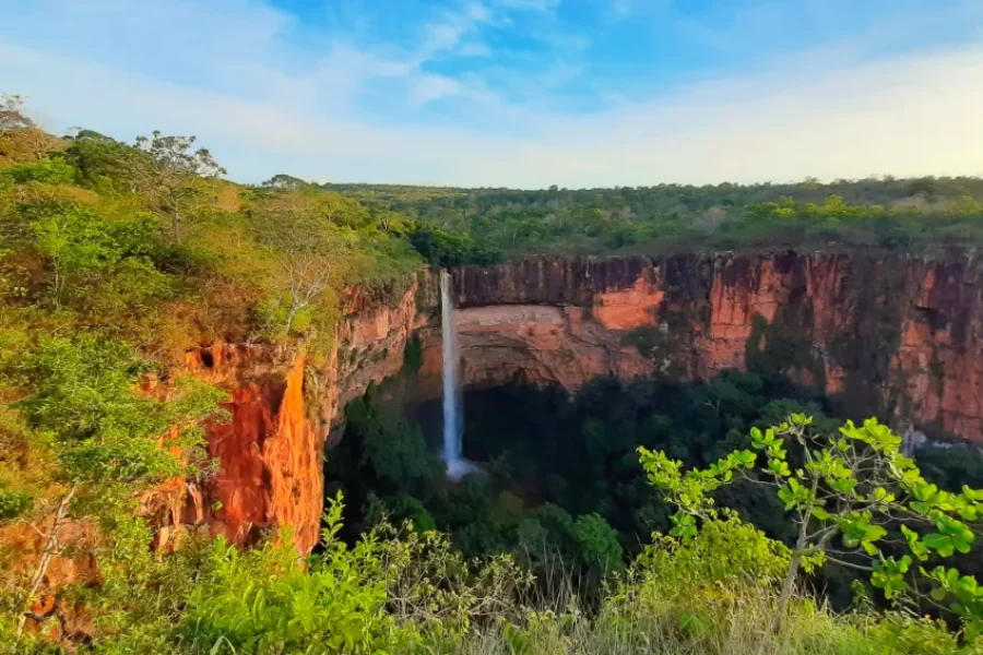 A waterfall plunging down a canyon with reddish rock walls in Chapada dos Guimarães, surrounded by lush Cerrado vegetation under a blue sky, in Mato Grosso, Brazil.