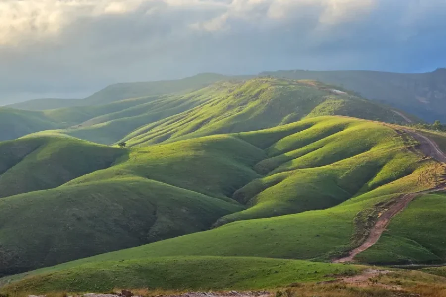Ondulações de campos verdes da Serra da Canastra com uma estrada de terra a serpentear pelas colinas sob céu nublado com raios de luz, no Parque Nacional da Serra da Canastra, Minas Gerais, Brasil.