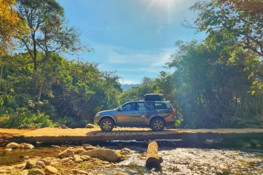 Jipe 4x4 a atravessar uma ponte de madeira rústica sobre um rio de águas cristalinas, rodeado de vegetação nativa do Cerrado sob céu azul, nos arredores de Delfinópolis, Minas Gerais, Brasil.