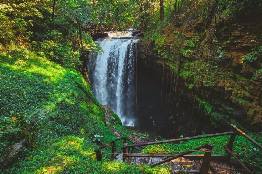 Cachoeira a despenhar-se entre paredes de rocha cobertas de vegetação exuberante do Cerrado, com escadas de madeira a conduzir ao miradouro, em Mambaí, Goiás, Brasil.