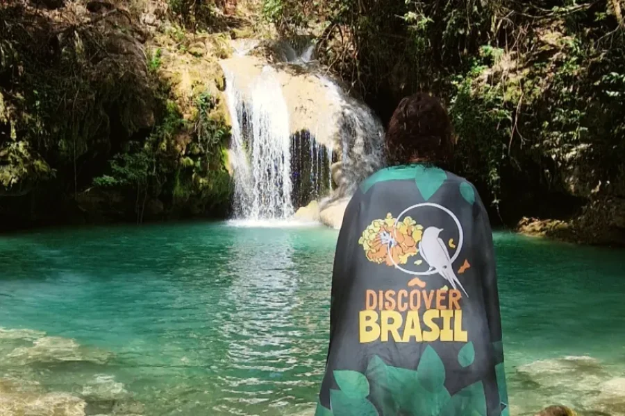Viajante de costas com bandeira da Discover Brasil contempla uma piscina natural de águas turquesa com cachoeira e vegetação de Cerrado ao redor, em Mambaí, Goiás, Brasil.