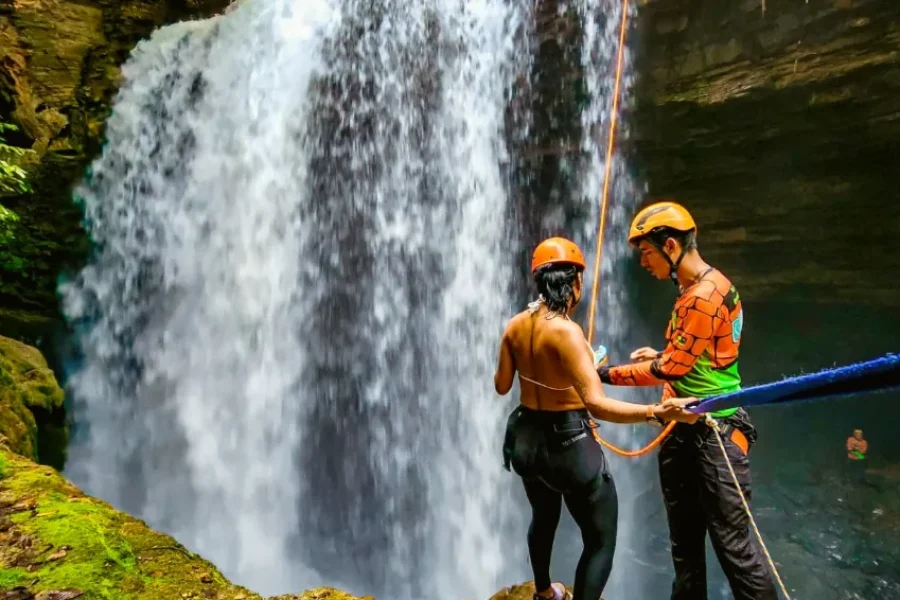 Guia a preparar viajante com equipamento de rapel em frente a uma cascata de pedra coberta de musgo verde, numa atividade de canyoning em Mambaí, Goiás, Brasil.