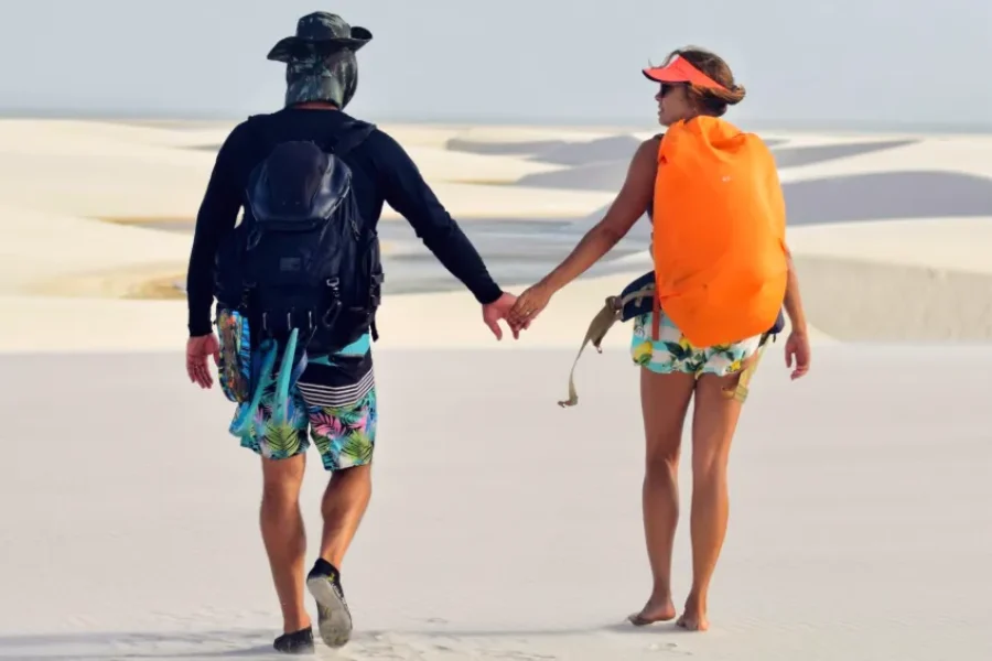 A couple holding hands with backpacks walking through the white dunes of Lençóis Maranhenses, on a trekking experience for two in the heart of Lençóis Maranhenses National Park, Maranhão, Brazil.