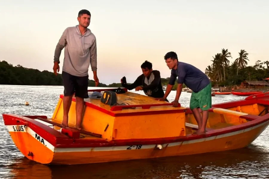 Três pescadores num barco de madeira alaranjado nas águas calmas do rio ao entardecer, com coqueiros e a vila de Barreirinhas ao fundo, no Maranhão, Brasil.