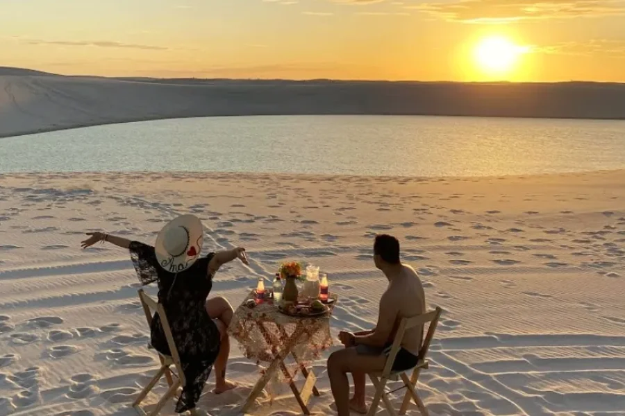 A couple sharing a romantic picnic at a table beside a lagoon, with the golden sunset reflecting off the white dunes of Lençóis Maranhenses, in Maranhão, Brazil.