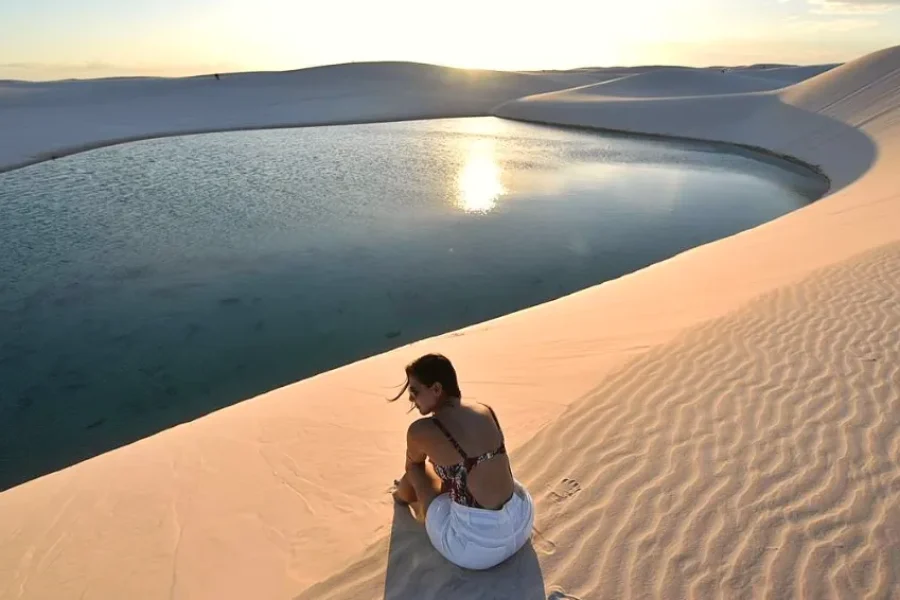 A traveller sitting on a white dune gazing at a turquoise lagoon at sunset, with the golden reflection of the sun on the water and the dunes of Lençóis Maranhenses stretching to the horizon, Maranhão, Brazil.