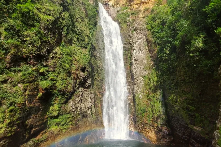 Cachoeira do Segredo precipitándose por una pared de roca encajada entre densa vegetación del Cerrado, con un arcoíris formándose en la base de la caída de agua, en la Chapada dos Veadeiros, Goiás, Brasil.