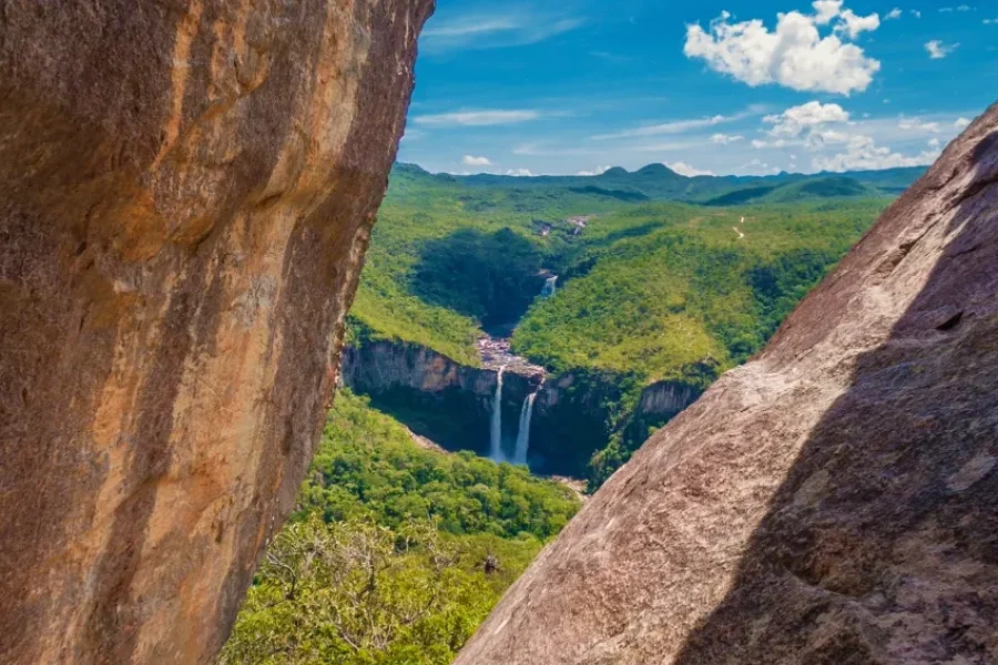 Vista enmarcada por dos formaciones rocosas en el Mirante da Janela, con la Cachoeira do Salto revelándose al fondo entre la exuberante vegetación del Cerrado y las sierras en el horizonte, en la Chapada dos Veadeiros, Goiás, Brasil.