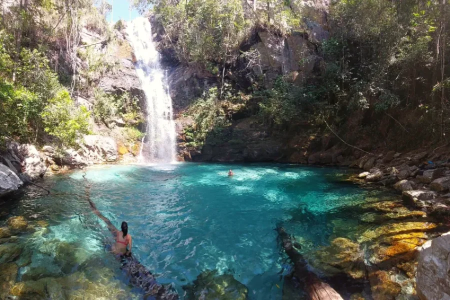 Bañistas en las aguas cristalinas y turquesas de la Cachoeira de Santa Bárbara, con la cascada precipitándose entre rocas y vegetación nativa del Cerrado, en Cavalcante, Goiás, Brasil.