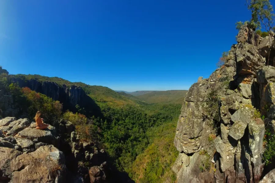 Viajero sentado en meditación en un mirador rocoso con vista panorámica sobre un valle cubierto de vegetación nativa del Cerrado y sierras en el horizonte bajo un cielo azul intenso, en la Chapada dos Veadeiros, Goiás, Brasil.