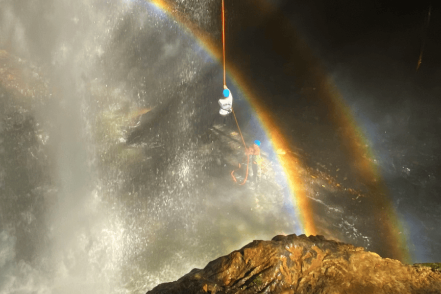 Person abseiling in Mambaí in front of a waterfall, with a rainbow formed by the mist, surrounded by the stunning natural scenery of Goiás.
