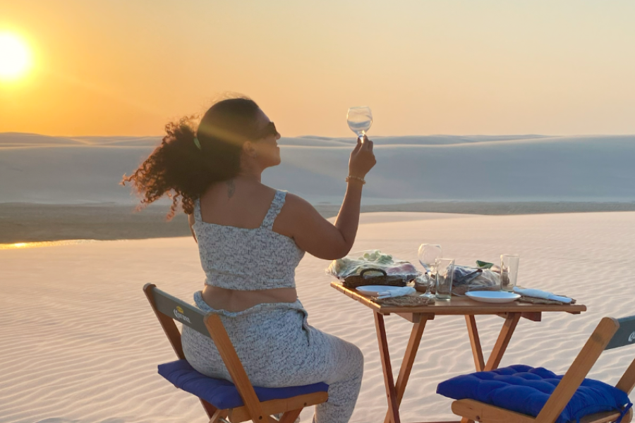 Woman toasting at sunset at a table set on the sand, overlooking the lagoons and dunes of Lençóis Maranhenses, near Barreirinhas.