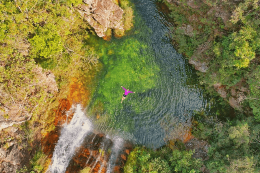 Person floating in a crystal-clear green pool beside a waterfall, surrounded by dense vegetation in the valleys of Delfinópolis.