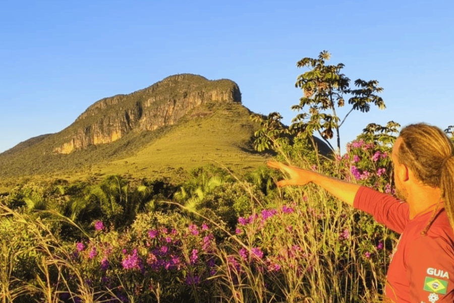 A local guide in Chapada dos Veadeiros points towards Whale Hill in the distance, framed by Cerrado vegetation and the flowers of the Maytrea Garden.