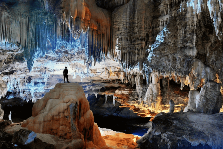 Explorer inside the Mambaí cave, lit by rock formations, stalactites, and bluish and orange hues.