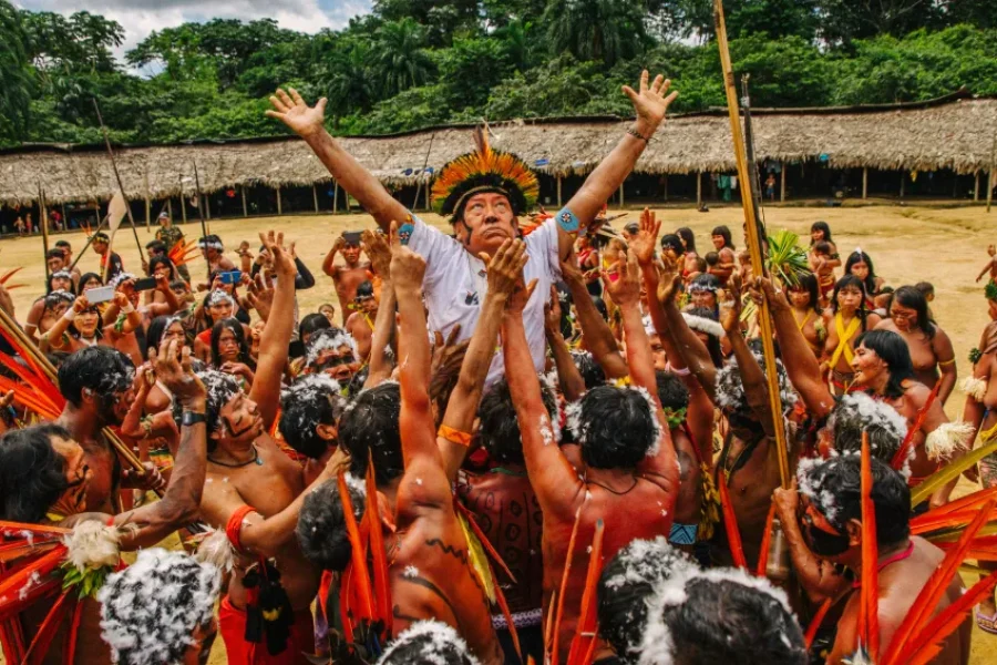 Eine gemeinschaftliche Feier des Yanomami-Volkes im zentralen Innenhof der Maloca, mit Gemeindemitgliedern in traditionellen Federschmuck und Körperbemalung, die die Arme zum Fest erheben, im Bundesstaat Amazonas, Brasilien.