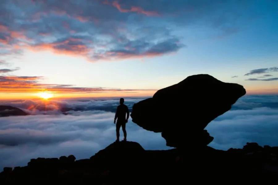 Silueta de un viajero en la cima del Monte Roraima al amanecer, con un mar de nubes abajo y el sol naciendo en tonos naranja y azul, en la frontera entre Brasil, Venezuela y Guyana.