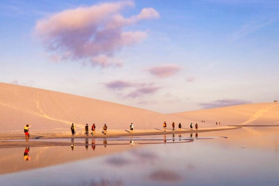 A group of travellers walking among dunes and a crystal-clear lagoon in the Lençóis Maranhenses at sunset, with the landscape reflected in the water.