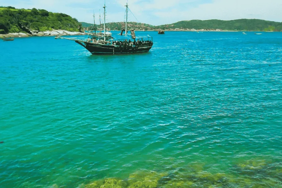 Eine hölzerne Schoner liegt in den kristallklaren, grünlichen Gewässern der Bucht von Búzios vor Anker, mit bewaldeten Hügeln im Hintergrund, in Armação dos Búzios, Rio de Janeiro, Brasilien.