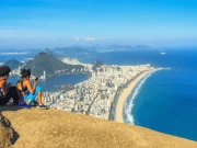 Due viaggiatrici sedute sulla cima del Morro Dois Irmãos che ammirano e fotografano la vista panoramica sulla spiaggia di Ipanema, la Lagoa Rodrigo de Freitas, il Pan di Zucchero e il lungomare di Rio de Janeiro, Brasile.