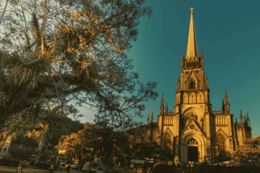 The Gothic façade of the Cathedral of São Pedro de Alcântara in Petrópolis, its pointed tower rising above lush vegetation under a blue sky, in the Serra Fluminense, Rio de Janeiro, Brazil.