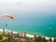 Un parapendio con vela arancione e gialla che sorvola la Barra da Tijuca, con una vista aerea sull'oceano Atlantico, la spiaggia e gli edifici lungo la costa di Rio de Janeiro, Brasile.