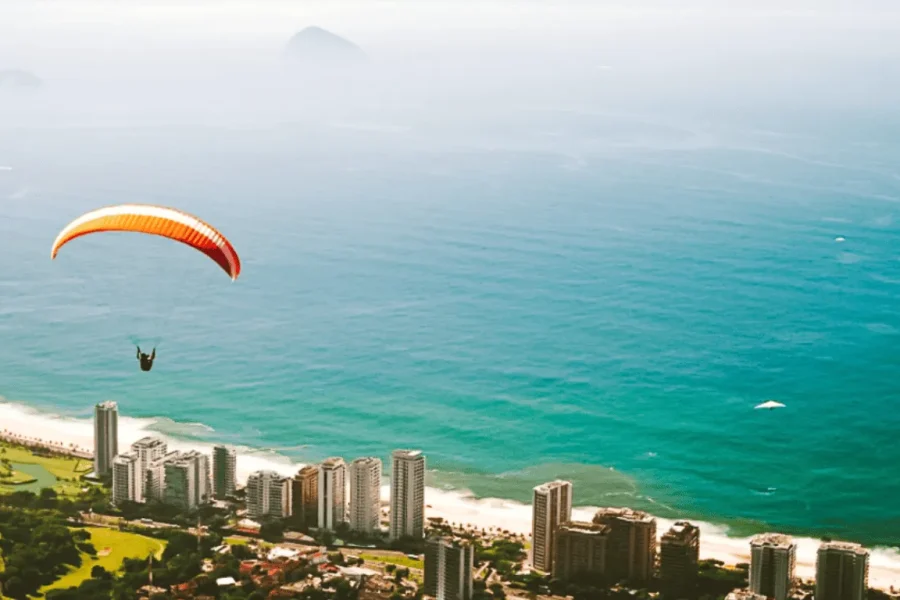 Ein Gleitschirm mit orangefarbener und gelber Kappe überfliegt die Barra da Tijuca, mit einem Luftblick auf den Atlantischen Ozean, den Strand und die Gebäude entlang der Küste von Rio de Janeiro, Brasilien.