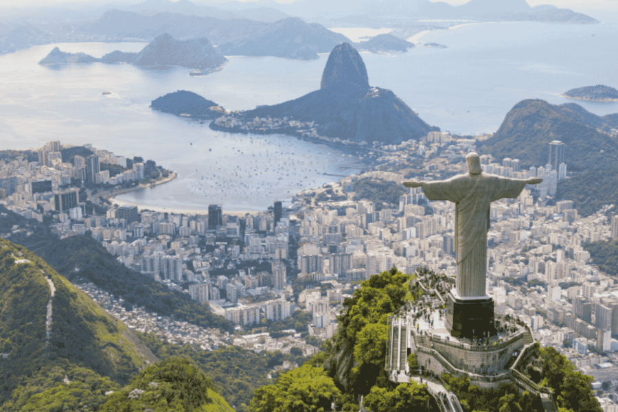 Aerial view of Christ the Redeemer with Guanabara Bay, Sugarloaf Mountain and the city of Rio de Janeiro in the background, surrounded by mountains and lush greenery.
