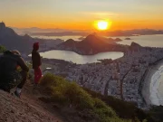 Due viaggiatori contemplano il tramonto dorato dalla cima del Morro Dois Irmãos, con una vista panoramica sulla Lagoa Rodrigo de Freitas, la spiaggia di Ipanema e la città di Rio de Janeiro, Brasile.