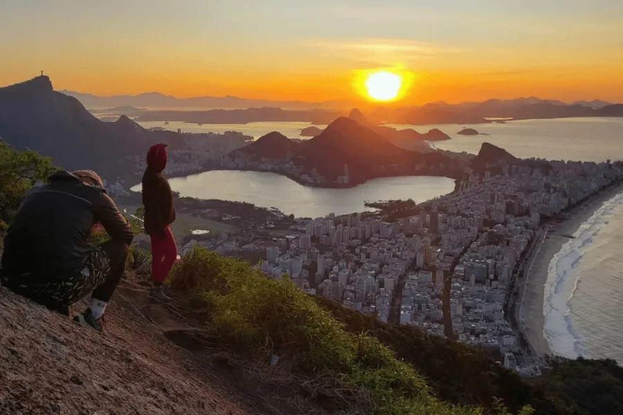 Two travellers gaze at the golden sunset from the top of Morro Dois Irmãos, with a panoramic view over Lagoa Rodrigo de Freitas, Ipanema beach and the city of Rio de Janeiro, Brazil.