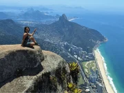 Una viaggiatrice seduta sulla cima della Pedra da Gávea che indica il paesaggio, con una vista mozzafiato sulla spiaggia di São Conrado, la Barra da Tijuca e il Morro Dois Irmãos sullo sfondo, a Rio de Janeiro, Brasile.
