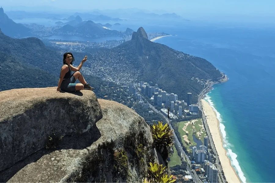 A traveller sitting at the summit of Pedra da Gávea pointing towards the landscape, with a breathtaking view over São Conrado beach, Barra da Tijuca and Morro Dois Irmãos in the background, in Rio de Janeiro, Brazil.