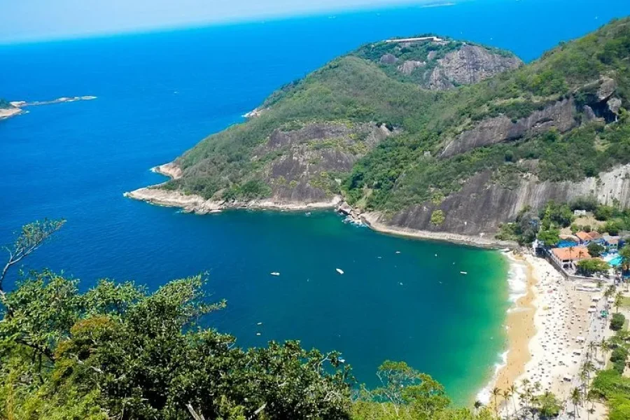 Luftaufnahme des Strandes Praia Vermelha und der Bucht des Morro da Urca, mit türkisfarbenem Wasser und goldenem Sand, umgeben von dichtem Atlantischen Regenwald und Felsenklippen, in Rio de Janeiro, Brasilien.