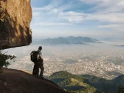 Un viaggiatore con zaino contempla la città di Rio de Janeiro avvolta nella nebbia da un belvedere roccioso al Bico do Papagaio, nella Foresta di Tijuca, con colline e la zona nord sullo sfondo, Rio de Janeiro, Brasile.