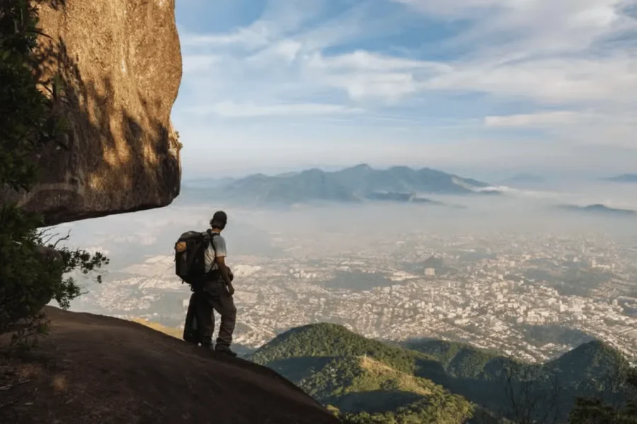 A backpack-wearing traveller gazes over the mist-covered city of Rio de Janeiro from a rocky viewpoint at Bico do Papagaio, in the Tijuca Forest, with hills and the north zone stretching into the distance, Rio de Janeiro, Brazil.