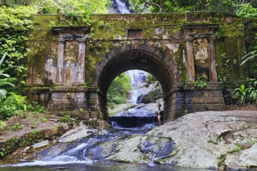 A historic moss-covered stone arch in the Tijuca Forest, with a waterfall flowing underneath and lush Atlantic Forest vegetation all around, in Rio de Janeiro, Brazil.