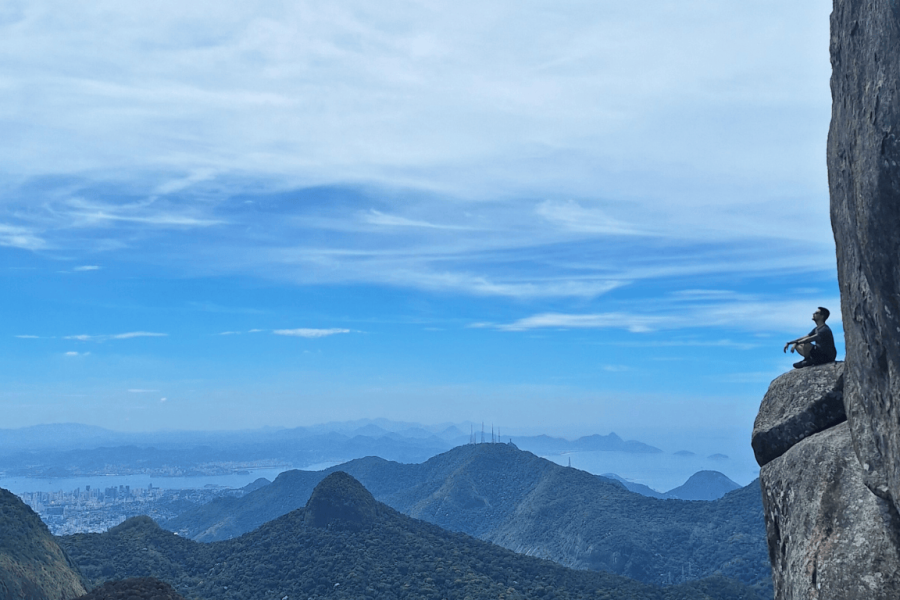 Person sitting on the edge of Bico do Papagaio in the Tijuca Forest, gazing at a wide view of the mountains and the city of Rio de Janeiro under a clear blue sky.