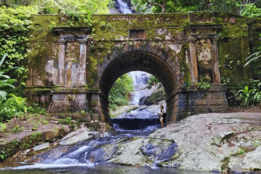 Woman standing under a stone arch with a waterfall in the background in the Tijuca Forest, surrounded by Atlantic Forest vegetation, during an eco-hike in Rio de Janeiro.