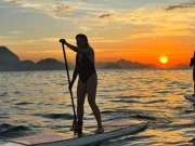 Due praticanti di stand-up paddle in silhouette sulle acque calme di Copacabana all'alba, con il sole che sorge in tonalità arancioni e il Pan di Zucchero sullo sfondo, a Rio de Janeiro, Brasile.