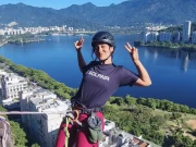 Una viaggiatrice sorridente con casco e attrezzatura da arrampicata che fa il segno della vittoria in un punto elevato sopra la Praia Vermelha, con la Lagoa Rodrigo de Freitas e la città di Rio de Janeiro sullo sfondo, Brasile.