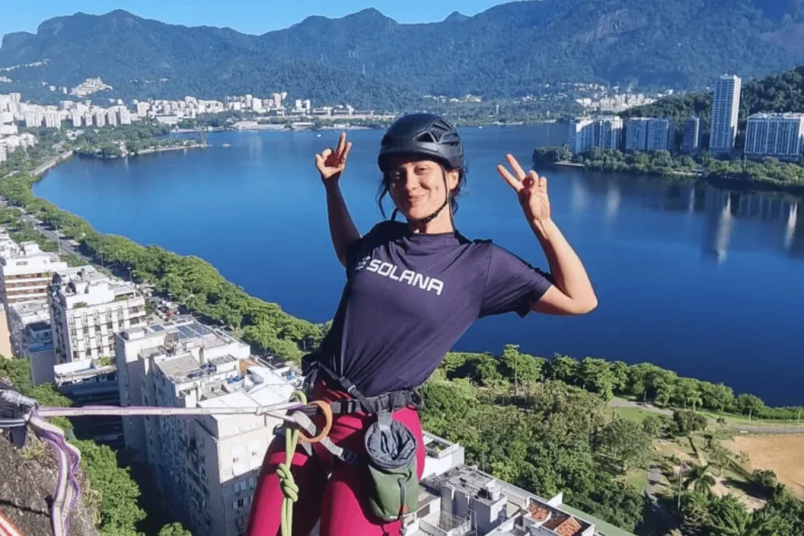 A smiling traveller wearing a helmet and climbing gear making a victory sign at an elevated point above Praia Vermelha, with Lagoa Rodrigo de Freitas and the city of Rio de Janeiro in the background, Brazil.