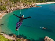 Un viaggiatore a braccia aperte che fa rappel su una scogliera sopra un'insenatura di acque turchesi e sabbia bianca, con la foresta atlantica sullo sfondo, su una spiaggia selvaggia di Rio de Janeiro, Brasile.