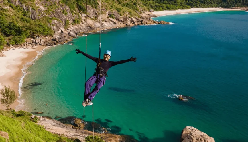Viajante de braços abertos a fazer rapel numa falésia sobre uma enseada de águas turquesa e areia branca, com a mata atlântica ao fundo, numa praia selvagem do Rio de Janeiro, Brasil.