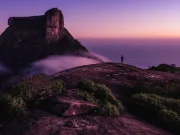 Silhouette di un viaggiatore in cima alla Pedra Bonita all'alba, con il cielo in tonalità lilla e rosa, la nebbia che avvolge le rocce e l'oceano Atlantico sullo sfondo, nella Foresta di Tijuca, Rio de Janeiro, Brasile.
