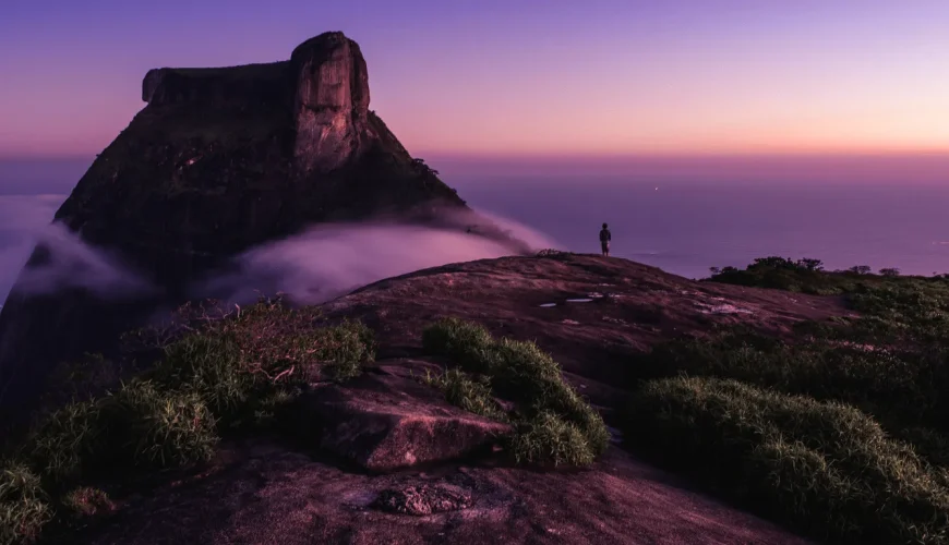 Silhueta de um viajante no cume da Pedra Bonita ao amanhecer, com o céu em tons de lilás e rosa, névoa a envolver as rochas e o oceano Atlântico ao fundo, na Floresta da Tijuca, Rio de Janeiro, Brasil.