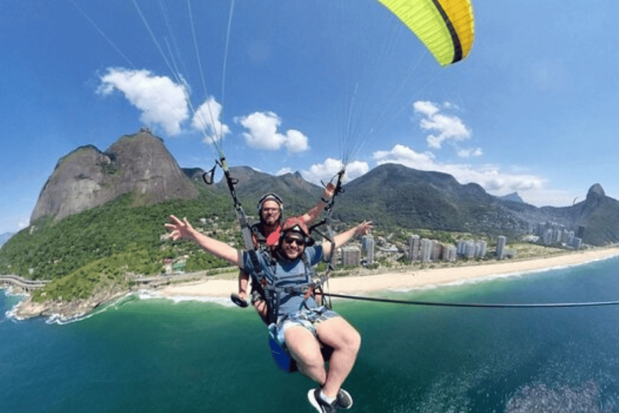 Two participants in a tandem paragliding flight over São Conrado Beach in Rio de Janeiro, with views of the sea, mountains, and Pedra da Gávea in the background.