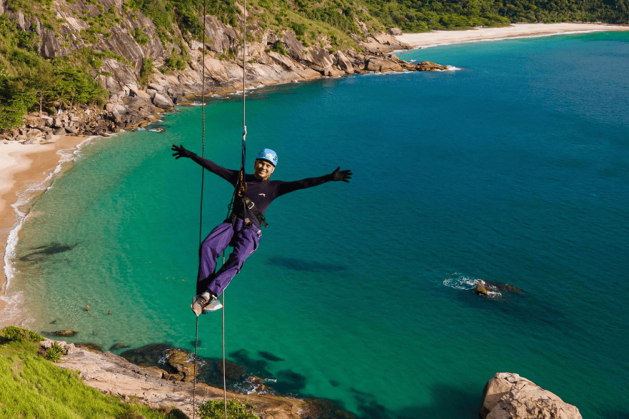 Person abseiling with safety equipment on a seaside rocky wall, overlooking a light sandy beach and blue sea in Rio de Janeiro.