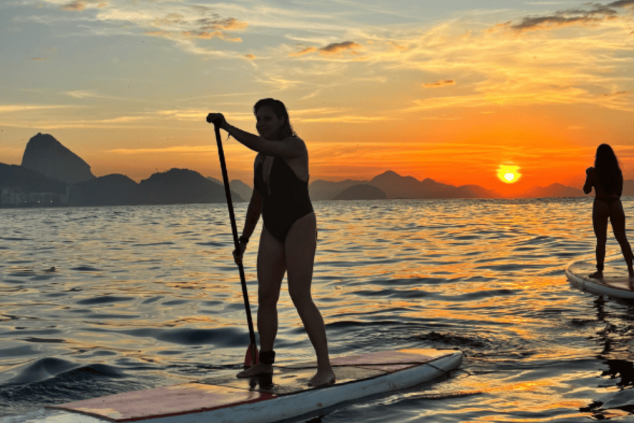 Person practising stand-up paddleboarding at sunrise in the calm waters of Copacabana Beach, with the sky painted by the rising sun in the background.