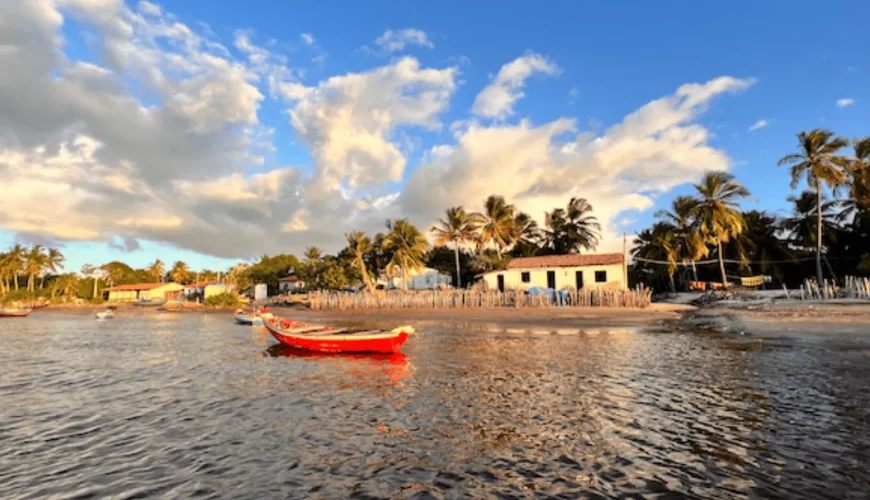 Barco de pesca vermelho ancorado nas águas calmas da praia de Atins, com casas de pescadores, coqueiros e céu nublado ao entardecer, no litoral do Maranhão, Brasil.